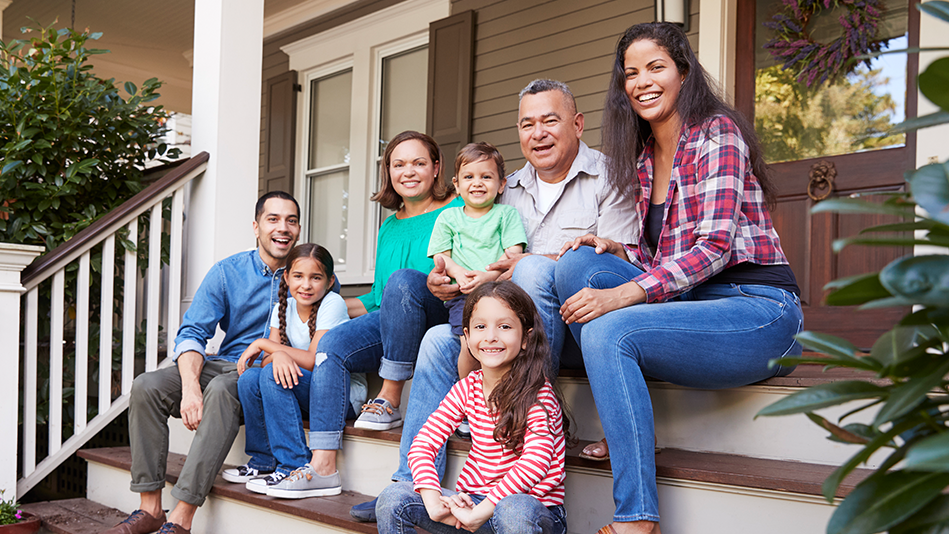Three generations of a Hispanic family smile while sitting on the steps in front of their home