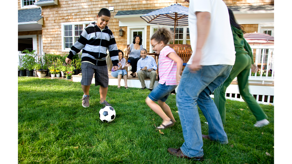 Three generations of a Hispanic family enjoy their yard on a nice  day. The oldest generation (2 women, 1 man) watch the two younger generations (1 man, 1 woman, 1 boy, 1 girl) play each  other in a game of soccer.