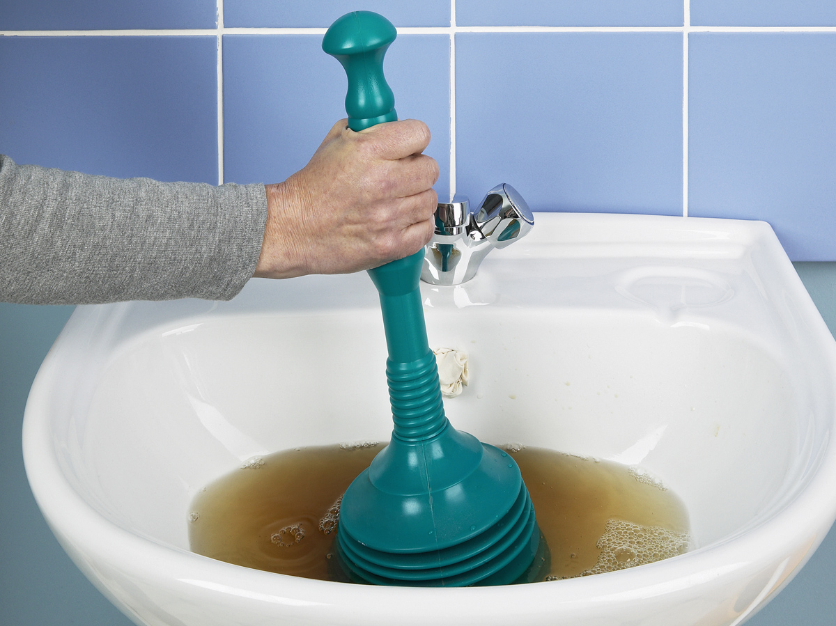 A white hand plunges brown water in a white bathroom sink mounted to a light blue-tiled wall 