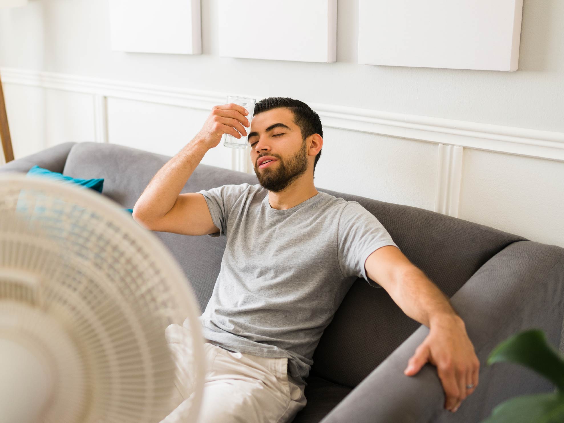 A Hispanic man puts a cool glass of water to his  head while sitting on the living room couch in front of a whirring fan