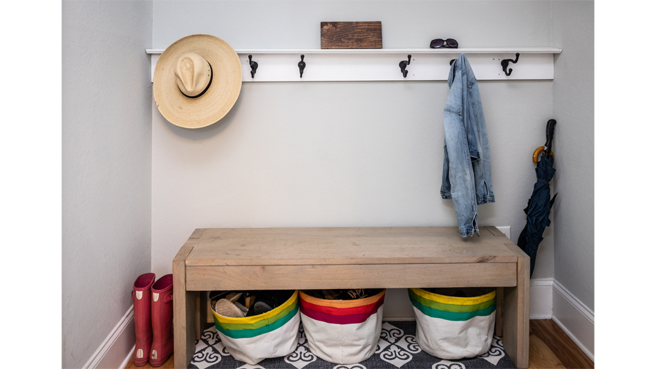 A small mudroom contains 6 hooks mounted to a white wood board, which is mounted to a white wall, a bench with multicolored canvas bucket baskets underneath; the baskets sit atop a white and gray/navy printed rug. A straw fedora and a jean jacket hang from the hooks; a black umbrella and a pair of hot pink rainboots flank the simple wooden bench. A small wooden plaque and a pair of sunglasses sit atop the wooden board the hooks hang from. The whole mudroom is in a recess in a hallway. 