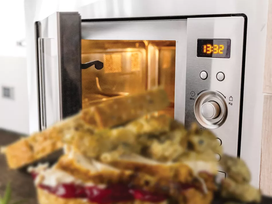 An open-faced rye bread sandwich with stuffing on top of sliced turkey on top of cranberry sauce in front of an open silver microwave with the clock reading "13:32"