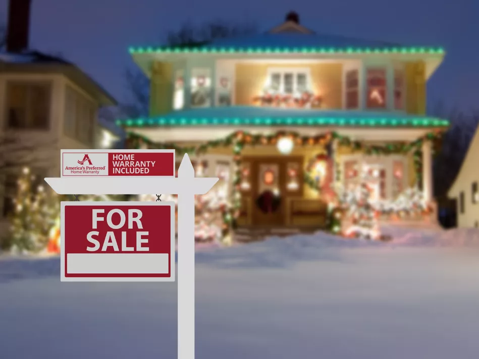 At night, a yellow two-story home is decorated and illuminated with holiday lights; the yard is covered with snow, and a red and white "For Sale" and APHW warranty rider sign are affixed to a white post in the front yard