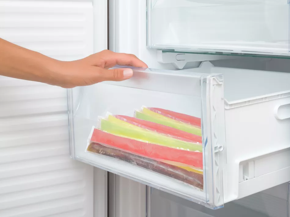 A light hand reaches for a stack of freezer pops in an open, transparent freezer drawer