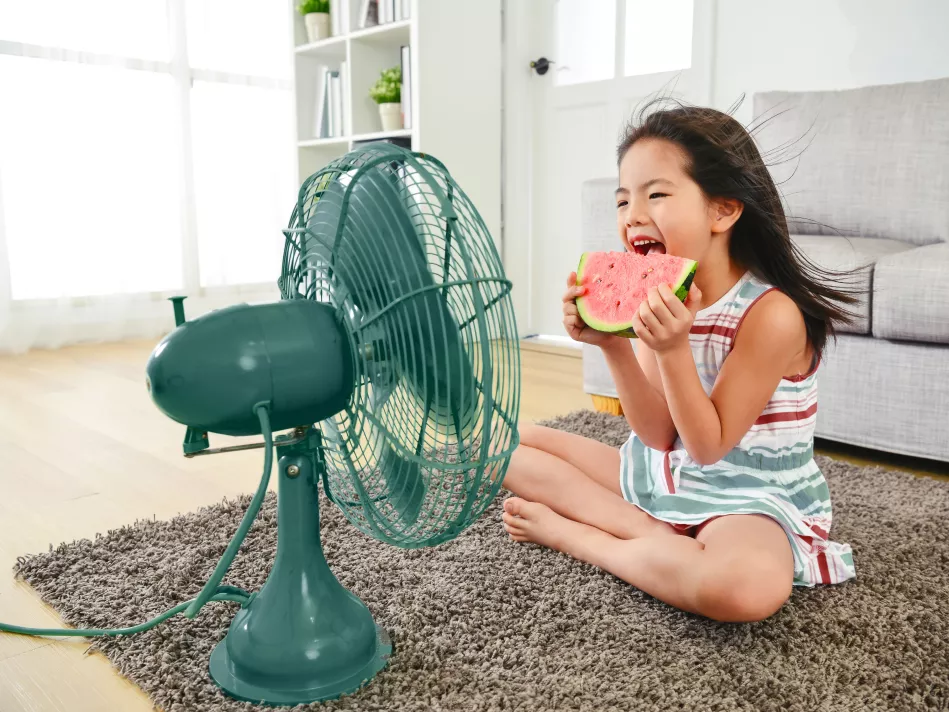 A little girl eats a slice of watermelon in front of a green fan 