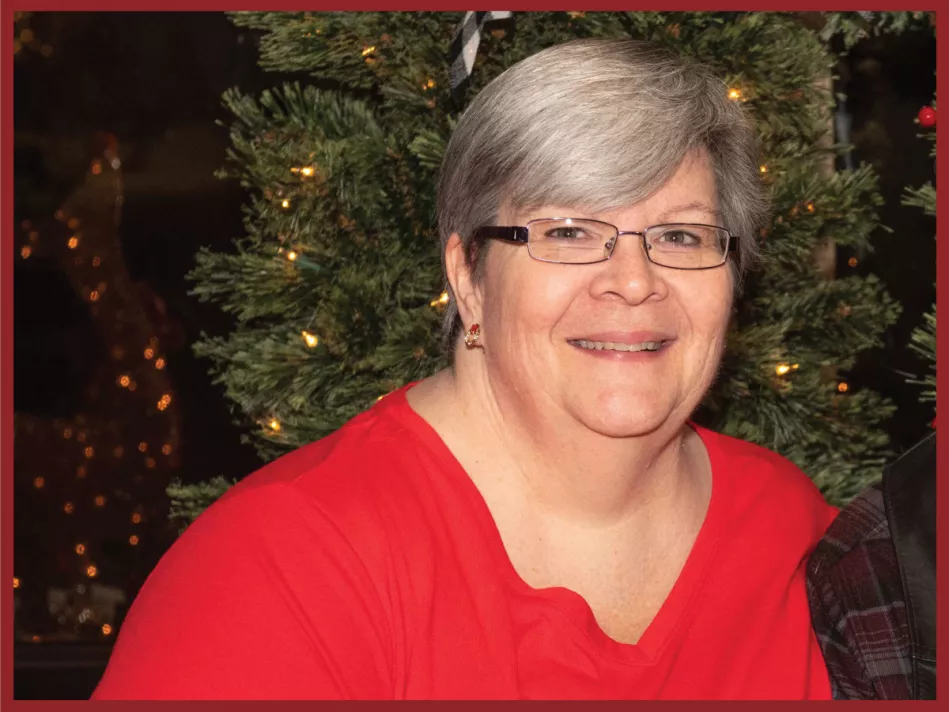 Kathy, a smiling grey haired woman wearing glasses and a red collared shirt in a holiday like scene is crowned as the Employee of the Month!