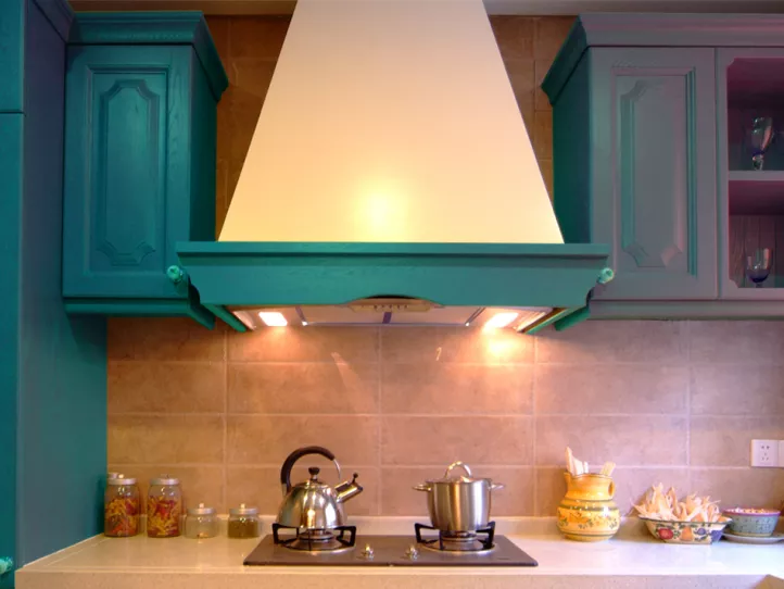 In a big, fancy kitchen, a silver kettle and small soup pot sit on each of the burners. The backsplash and counters are tan, and the cupboards are teal. Dry pasta and spices in various containers flank the stove itself.  