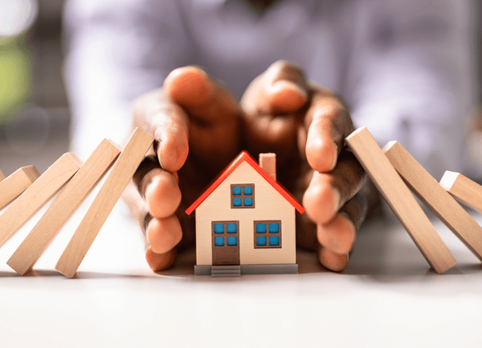 Hands protecting a miniature house from falling wooden blocks, symbolizing home protection, insurance, or risk management.