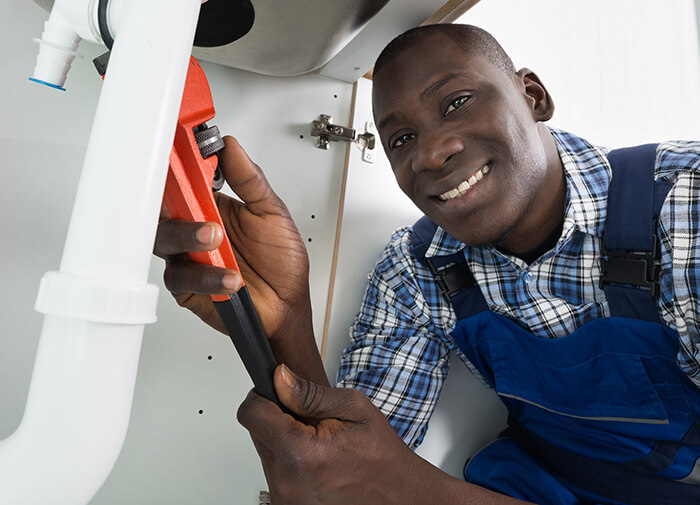 A smiling professional african american plumber using a wrench to fix pipes under a sink, wearing blue overalls and working confidently.