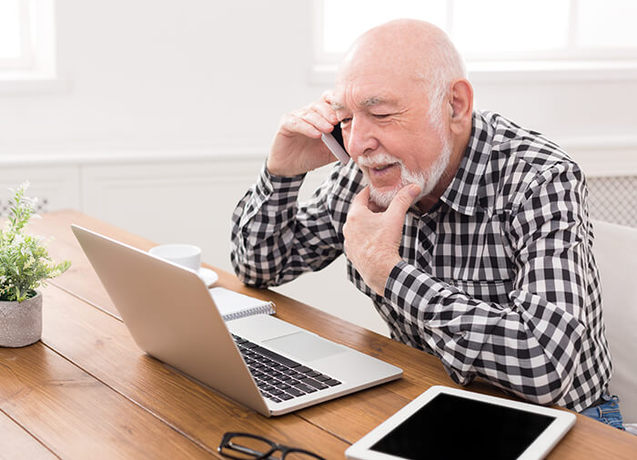 Senior man in a checkered shirt talking on the phone while using a laptop and tablet, working from home or consulting remotely.