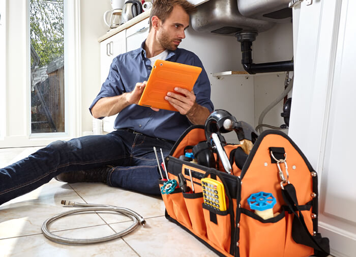 Professional plumber sitting on the floor using a tablet to troubleshoot a sink issue, with an orange tool bag and plumbing equipment beside him.