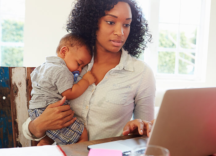 Working mother holding her sleepy baby while using a laptop, balancing childcare and remote work at home.